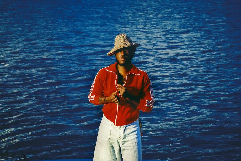 Soul musician Leon Bridges wearing a straw hat and red jacket stands outdoors in front of shimmering deep blue water.